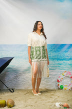 A woman stands on a beach-themed studio backdrop wearing a white short-sleeve collared dress with Naina Fringe Skirt | Green Botanical Resort Skirt with Fringe Hem. She is holding a small metallic clutch, with a black umbrella, coconuts, and a pink swim ring visible in the scene.