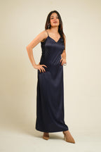 A woman modeling a navy blue satin slip dress with thin spaghetti straps and a V-neckline, standing against a plain beige studio background.