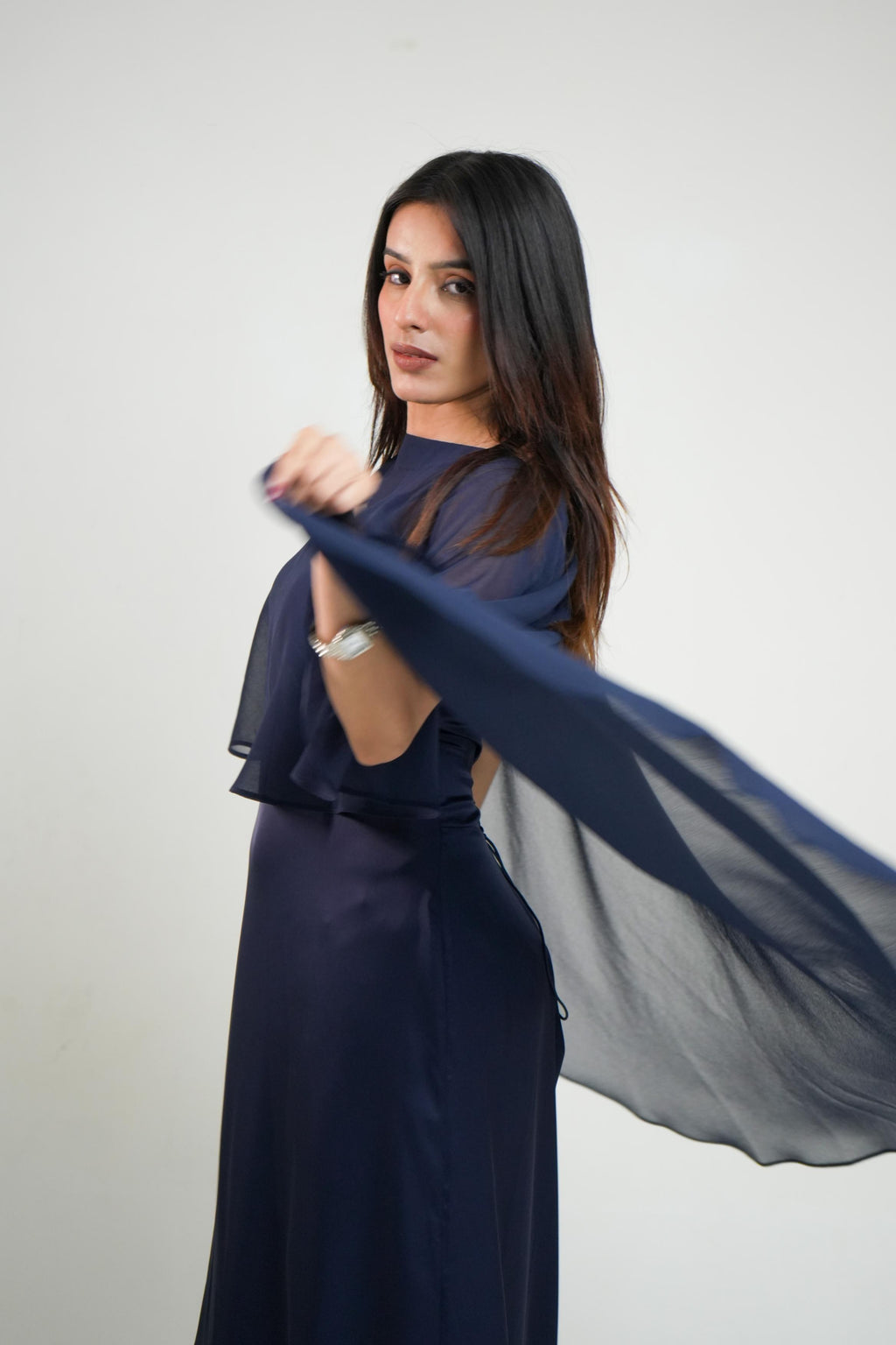 A woman modeling Nancy Navy Satin Cape Dress | Draped Occasion Dress for Women  a navy blue dress with a matching sheer fabric draped across her body, set against a plain light gray studio background.