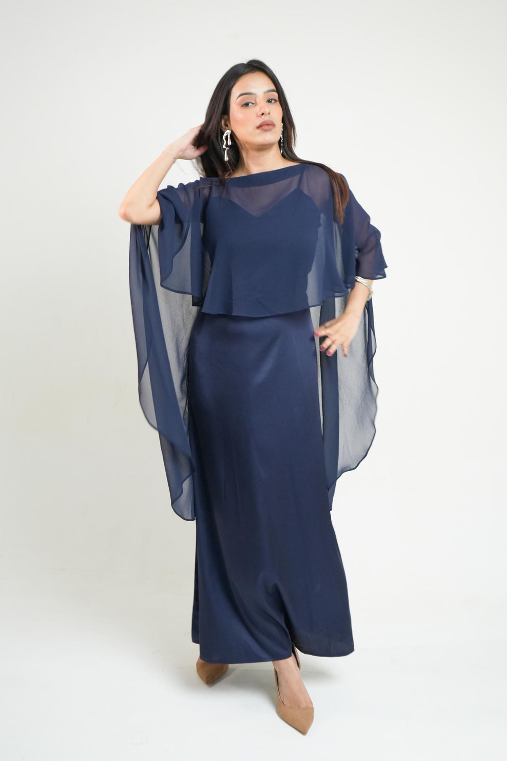 A woman modeling a navy blue two-piece dress set featuring a sheer mesh cape top and a matching longdress known as khamna Nancy Navy Satin Cape Dress | Draped Occasion Dress for Women against a plain light gray studio background.