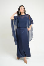 A woman modeling a navy blue two-piece dress set featuring a sheer mesh cape top and a matching longdress known as khamna Nancy Navy Satin Cape Dress | Draped Occasion Dress for Women against a plain light gray studio background.