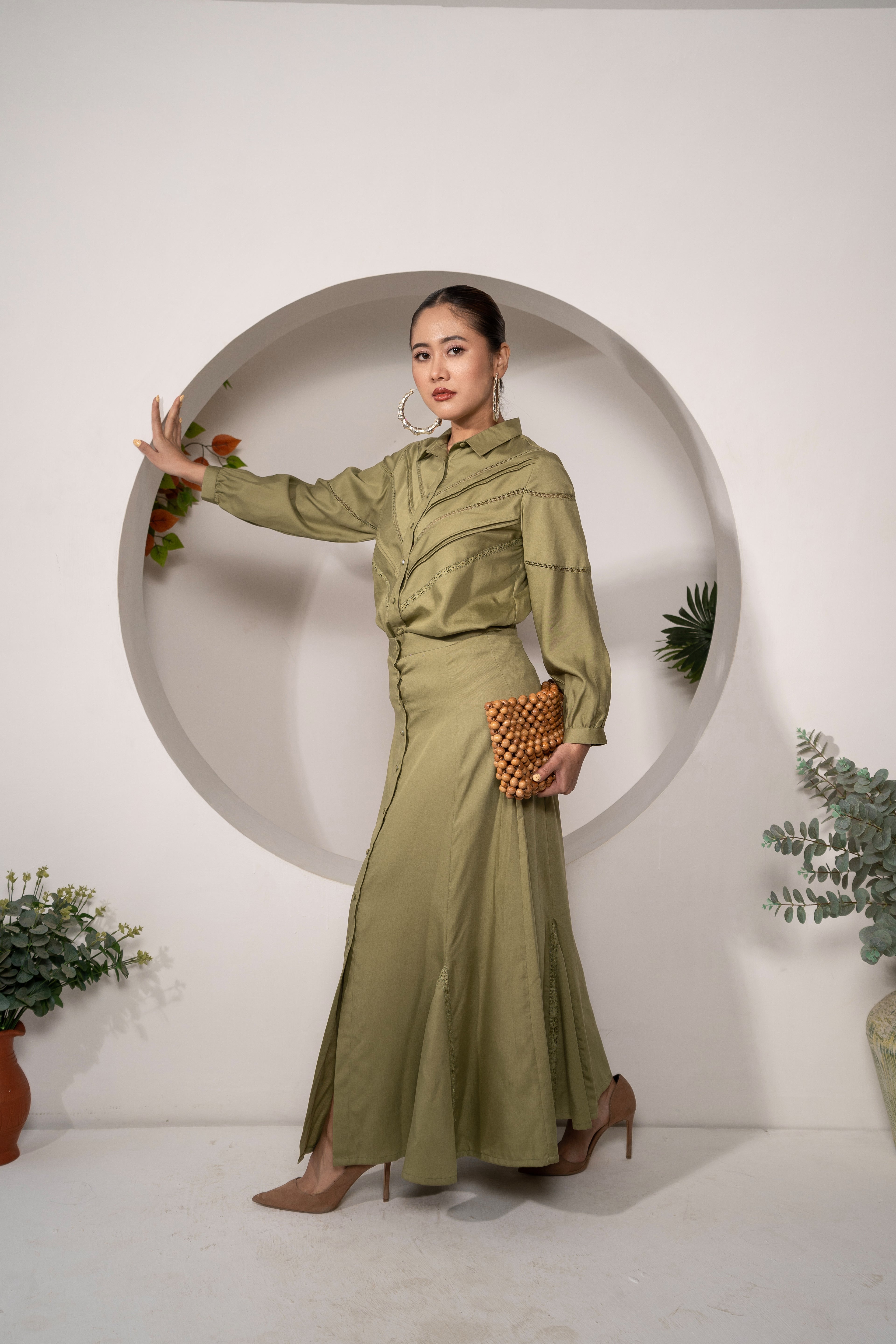 Woman in a Olive Reverie – Lace Detail Modal Shirt with matching skirt holding a clutch against a white wall with circular cutout