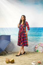 A woman modeling a red, blue, and black plaid midi dress with three-quarter sleeves, standing on a sandy beach backdrop with a blue umbrella, coconuts, and a swim ring nearby.