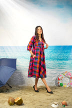 A woman modeling a colorful plaid midi dress with three-quarter sleeves, standing on a sandy beach backdrop with an ocean and sky background. She is wearing black pointed-toe heels.