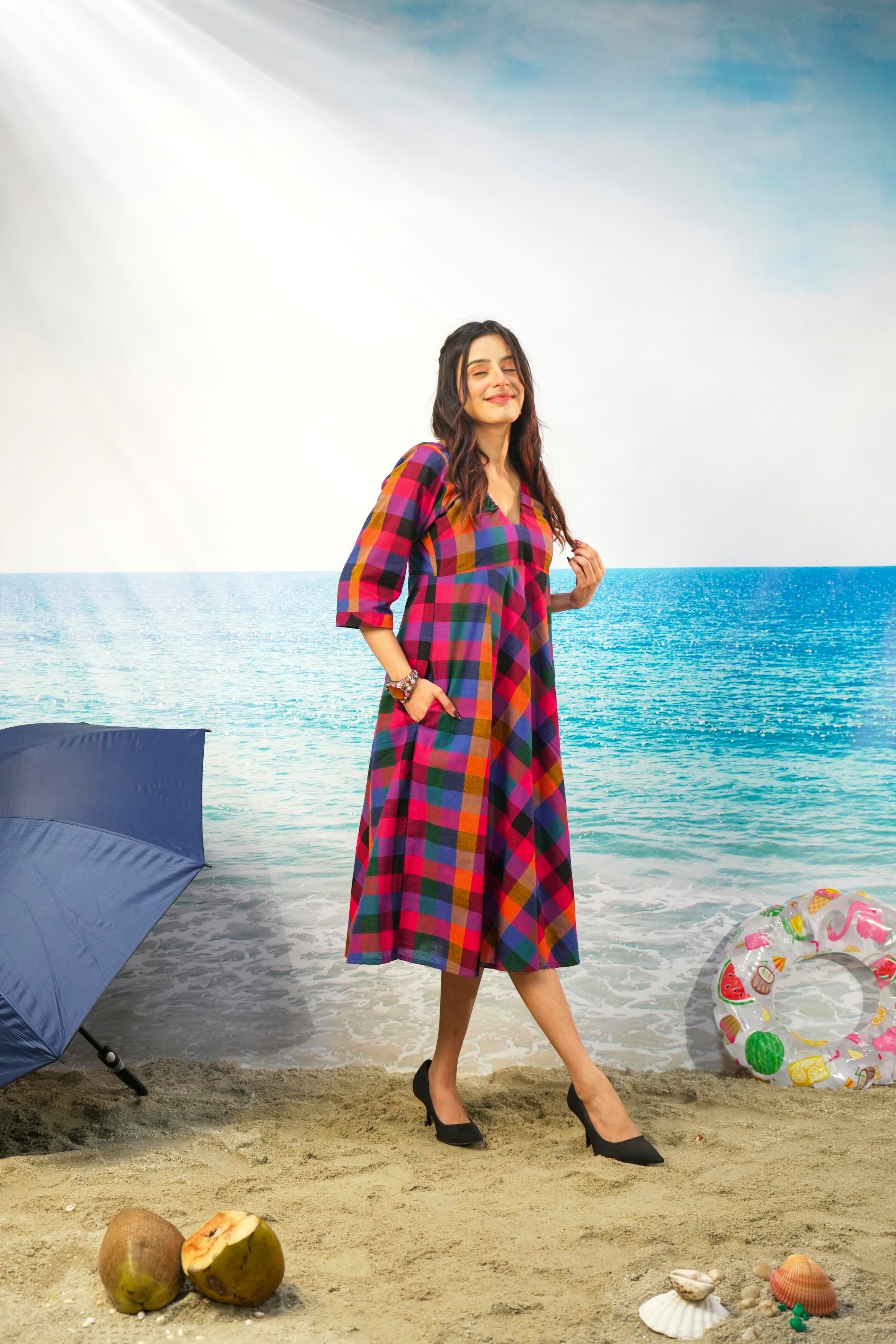 A woman modeling a colorful plaid midi dress with three-quarter sleeves, standing on a sandy beach backdrop with an ocean and sky background. She is wearing black pointed-toe heels.