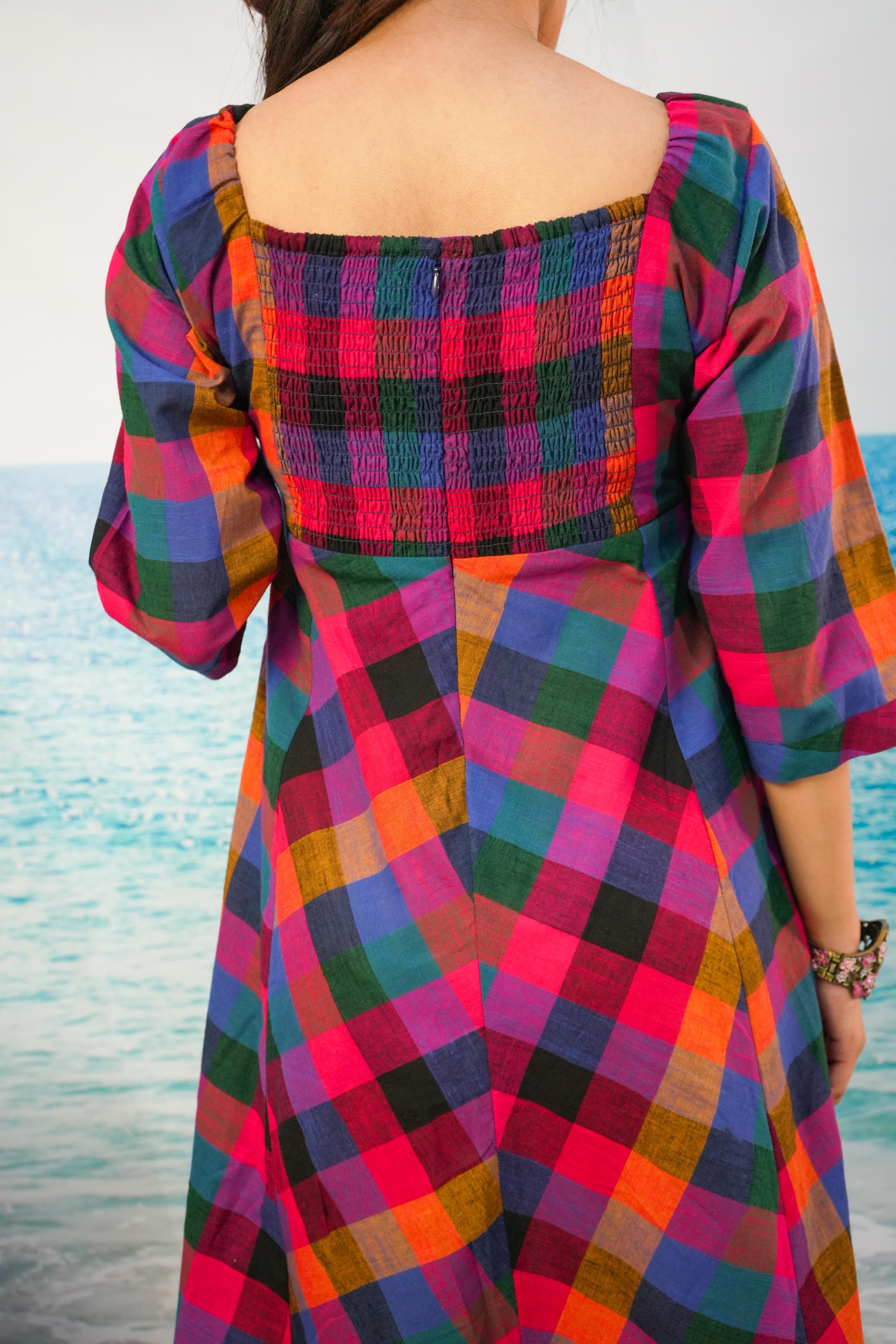 A woman viewed from the back wearing a colorful plaid dress with puff sleeves and a smocked yoke, set against a blurred beach and ocean background.