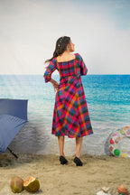 A woman stands on a beach backdrop wearing a colorful plaid midi dress with long sleeves and a square neckline. The dress has a bold pattern of pink, blue, yellow, and red.