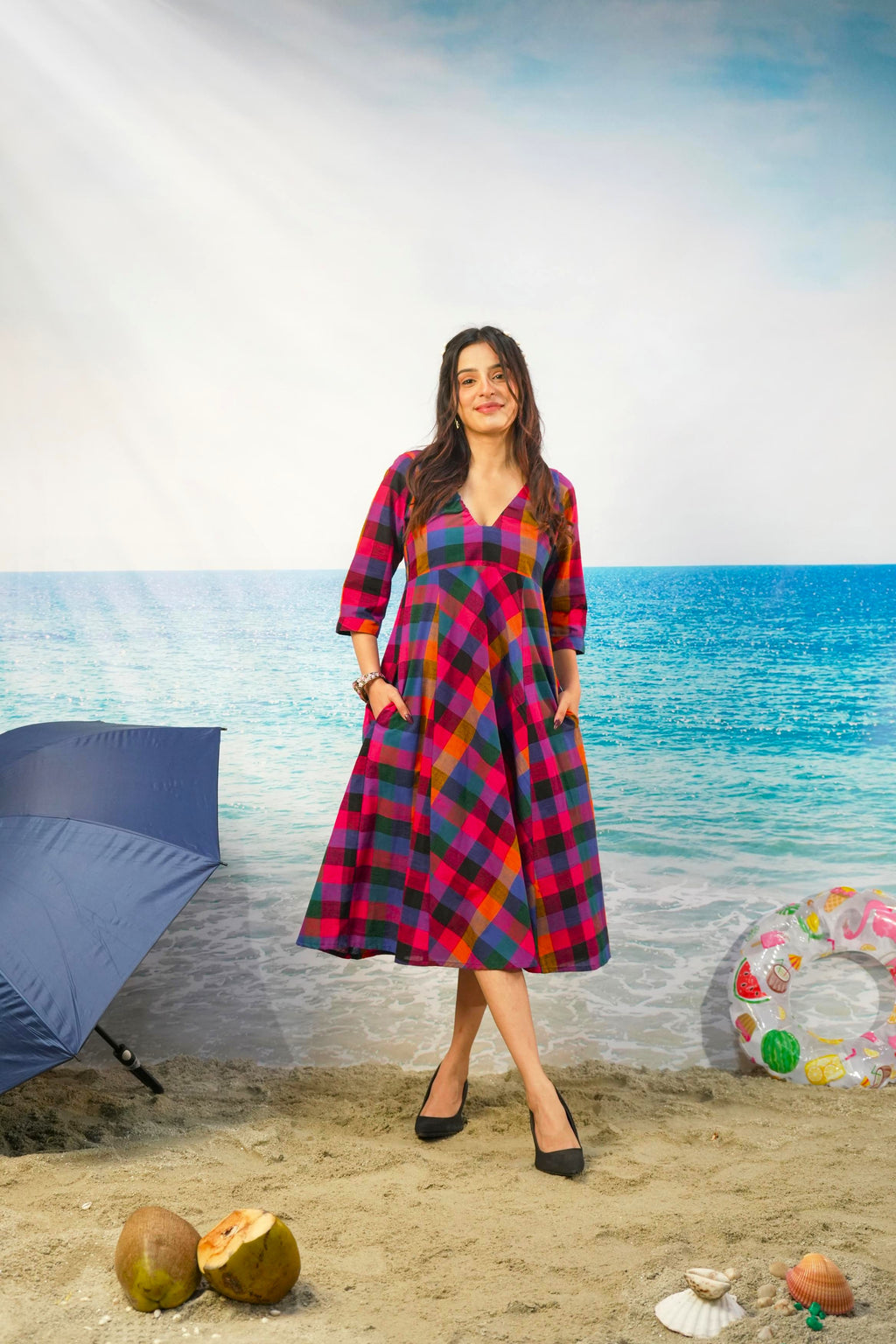 A woman modeling a red, blue, and yellow plaid midi dress with pockets, standing on a sandy beach backdrop with a blue umbrella, seashells, and a pool float nearby.