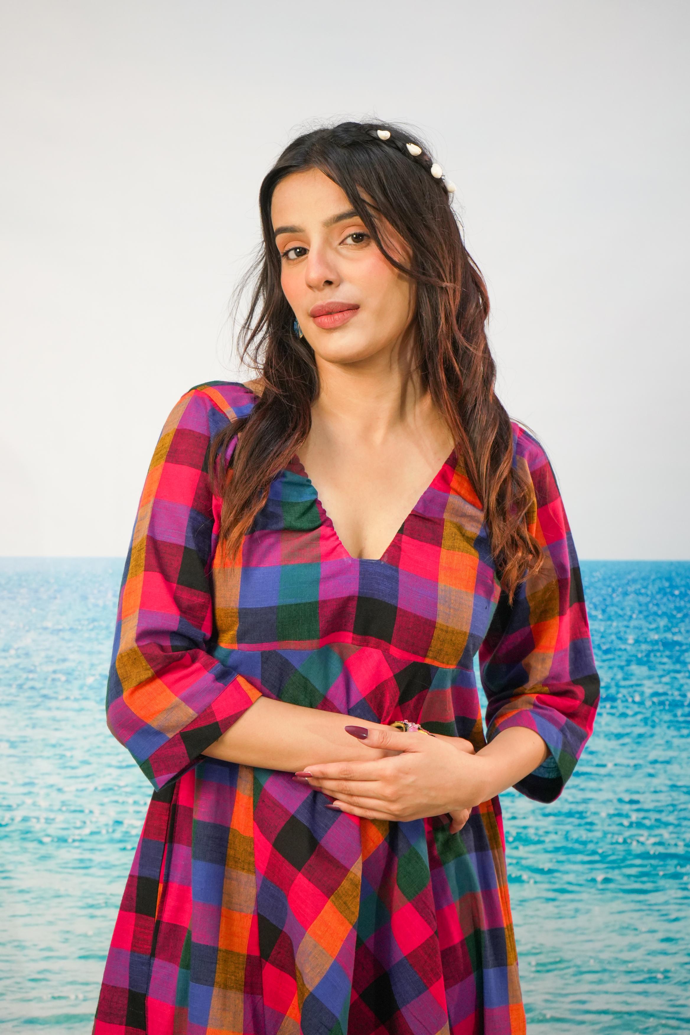 A woman with long dark wavy hair wearing a colorful plaid dress stands in front of a bright blue ocean under a pale sky. The dress has a vibrant multicolor checkered pattern with pink, orange, blue, green, and purple tones.
