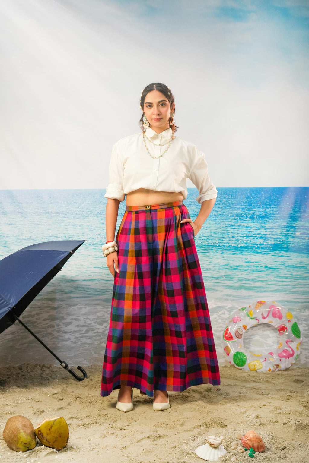 A woman stands on a sandy beach backdrop wearing a white cropped blouse and Ridhi Pleated Pants | Multicolor Handloom Madras Check Cotton Wide Leg Pants. A black beach umbrella, coconuts, shells, and a colorful float are visible around her.