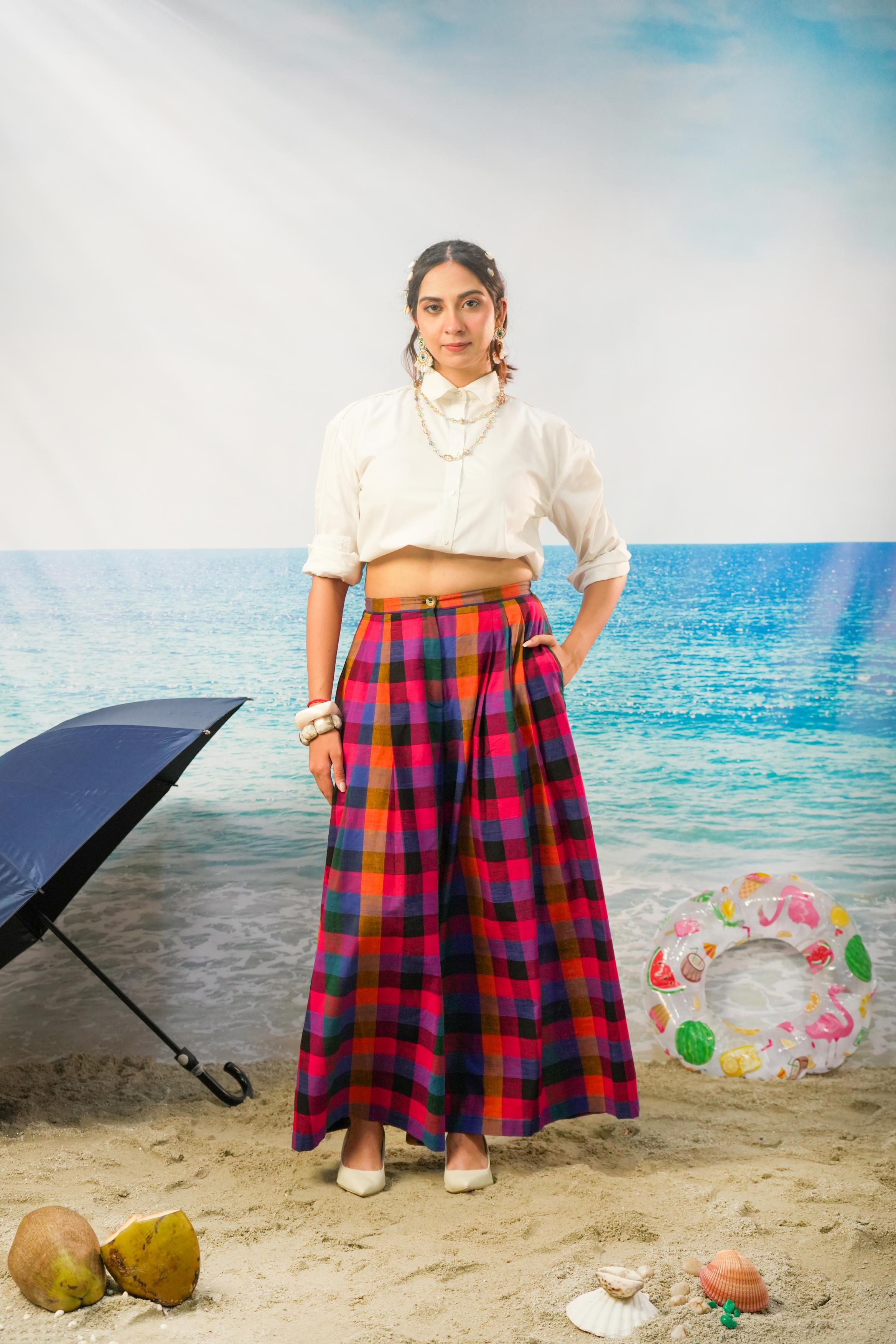 A woman stands on a sandy beach backdrop wearing a white cropped blouse and Ridhi Pleated Pants | Multicolor Handloom Madras Check Cotton Wide Leg Pants. A black beach umbrella, coconuts, shells, and a colorful float are visible around her.