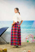 A woman stands on a beach backdrop wearing a white short-sleeve blouse and a long, wide-leg plaid skirt in pink, red, blue, and green. A black umbrella, coconuts, and a colorful inflatable ring float in the scene.