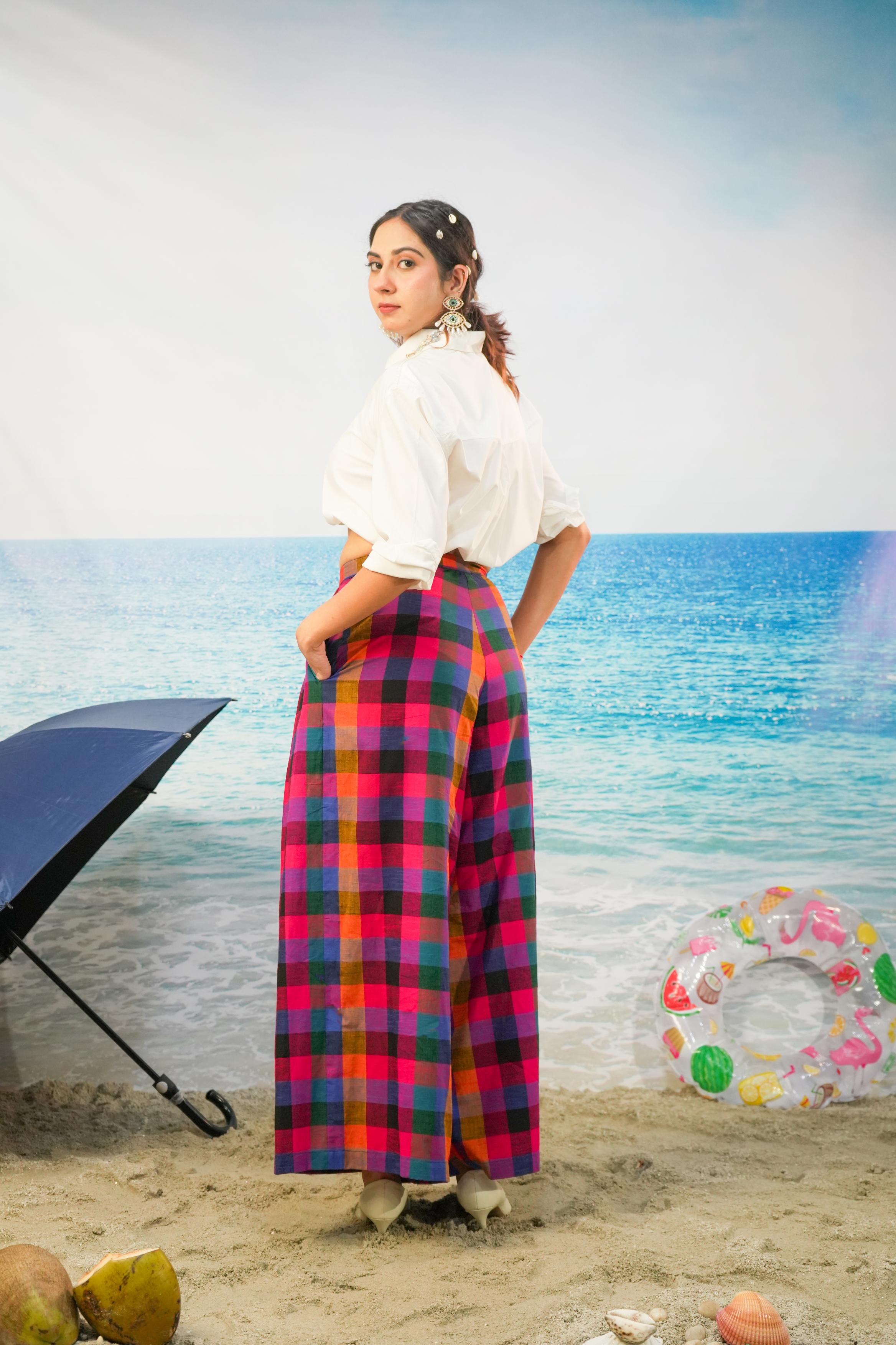 A woman stands on a beach backdrop wearing a white short-sleeve blouse and a long, wide-leg plaid skirt in pink, red, blue, and green. A black umbrella, coconuts, and a colorful inflatable ring float in the scene.