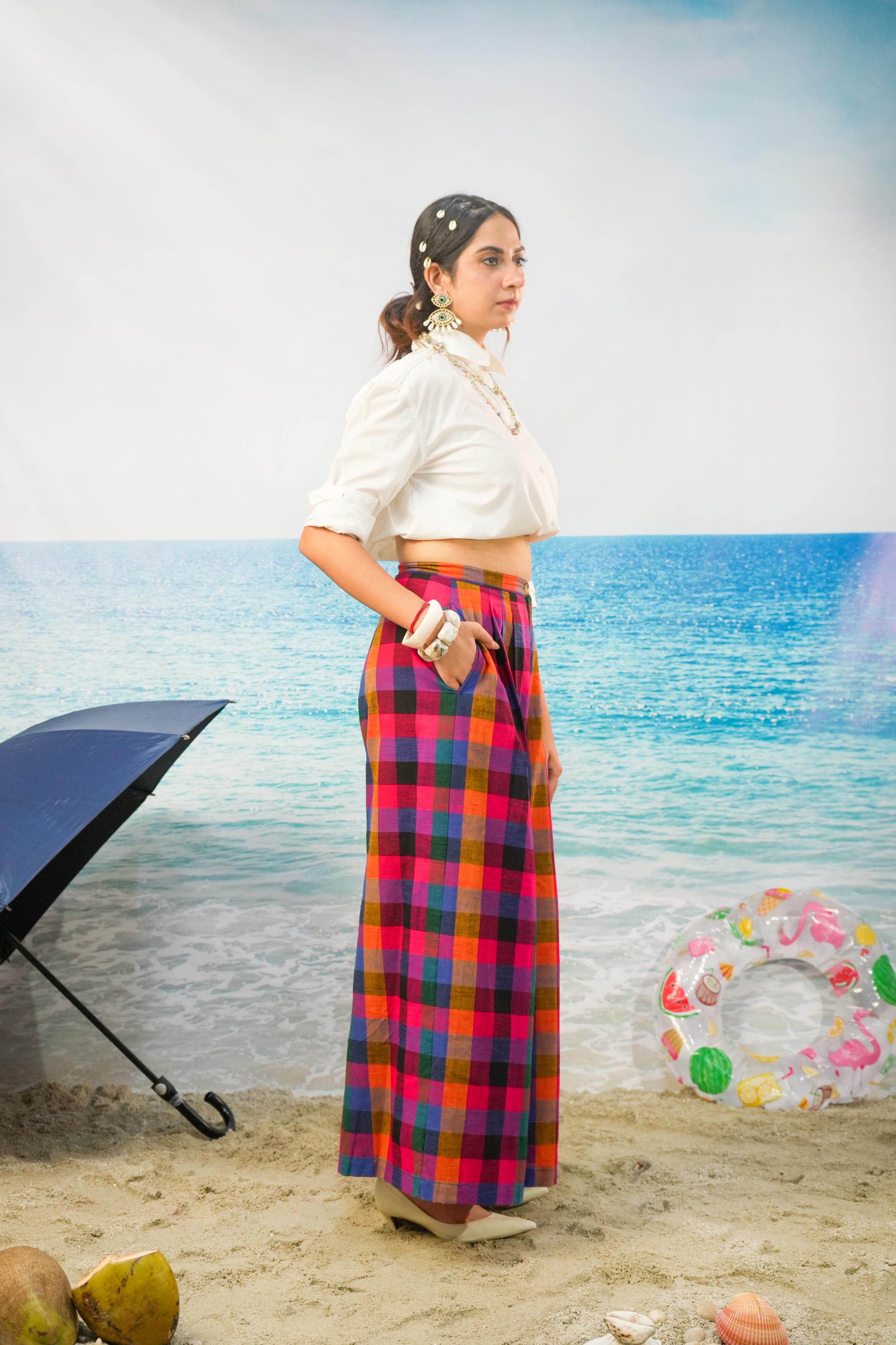 A woman stands on a sandy beach-like set wearing a white cropped blouse and a long multicolor plaid Pants. The background shows blue water, a pale sky, an open black umbrella, and a colorful inflatable ring.