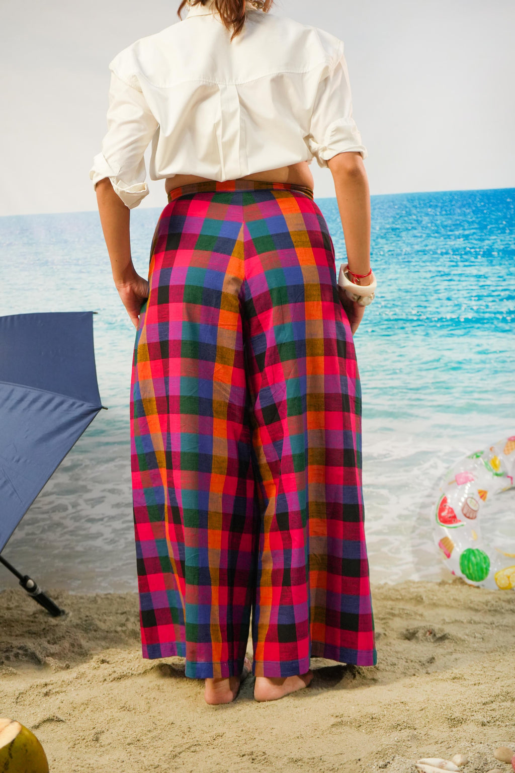 A person standing on a sandy beach facing the ocean, wearing bright multicolor plaid wide-leg pants and a white cropped blouse.