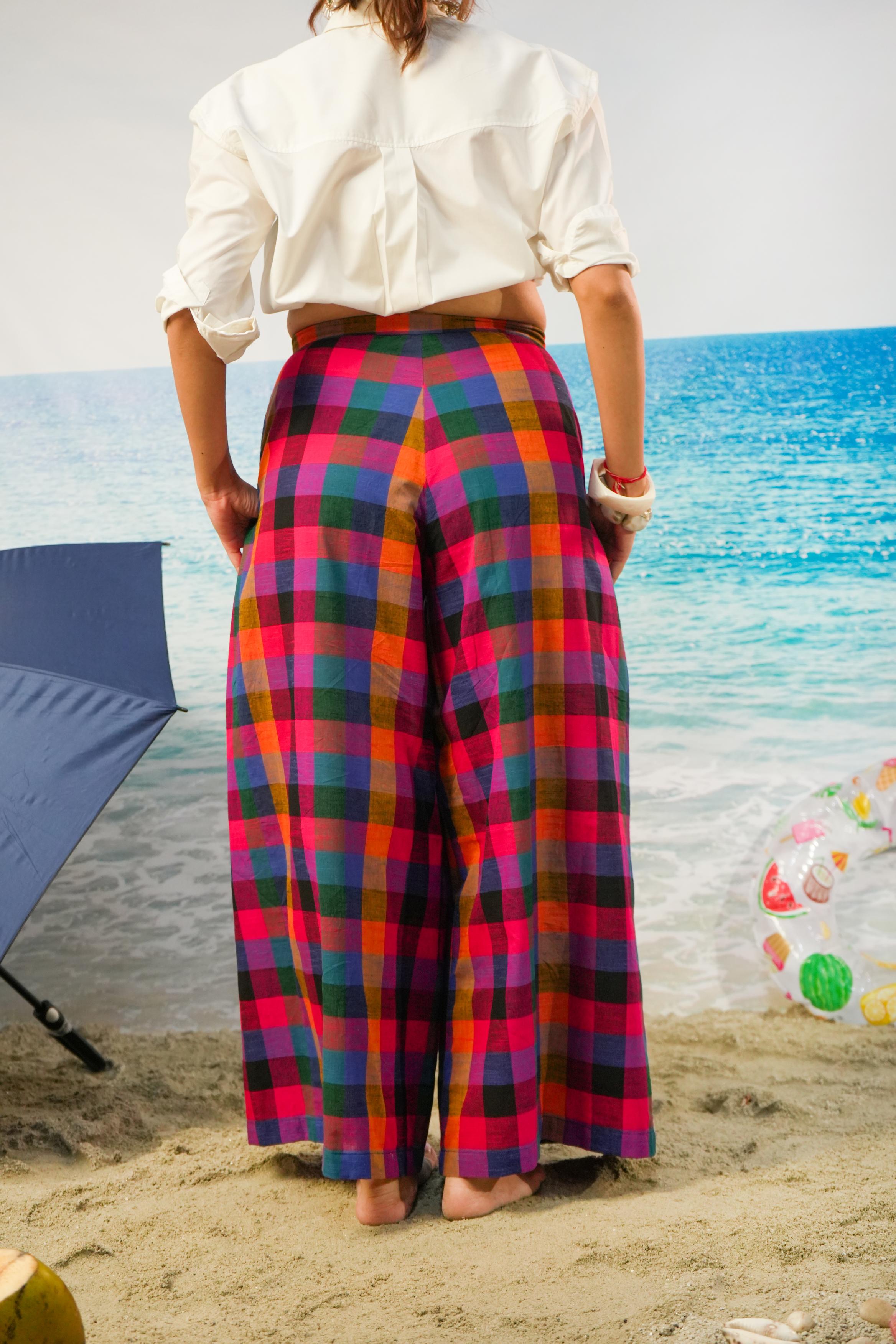 A person standing on a sandy beach facing the ocean, wearing bright multicolor plaid wide-leg pants and a white cropped blouse.