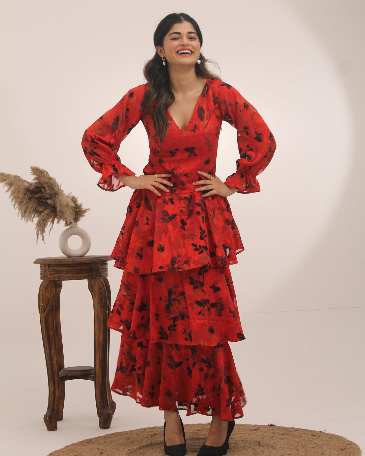 Woman in a red dress with black floral pattern posing against a white wall.