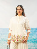 A woman modeling a cream-colored embroidered Blouse and matching trousers, set against a backdrop of a blue ocean and a light gray sky.