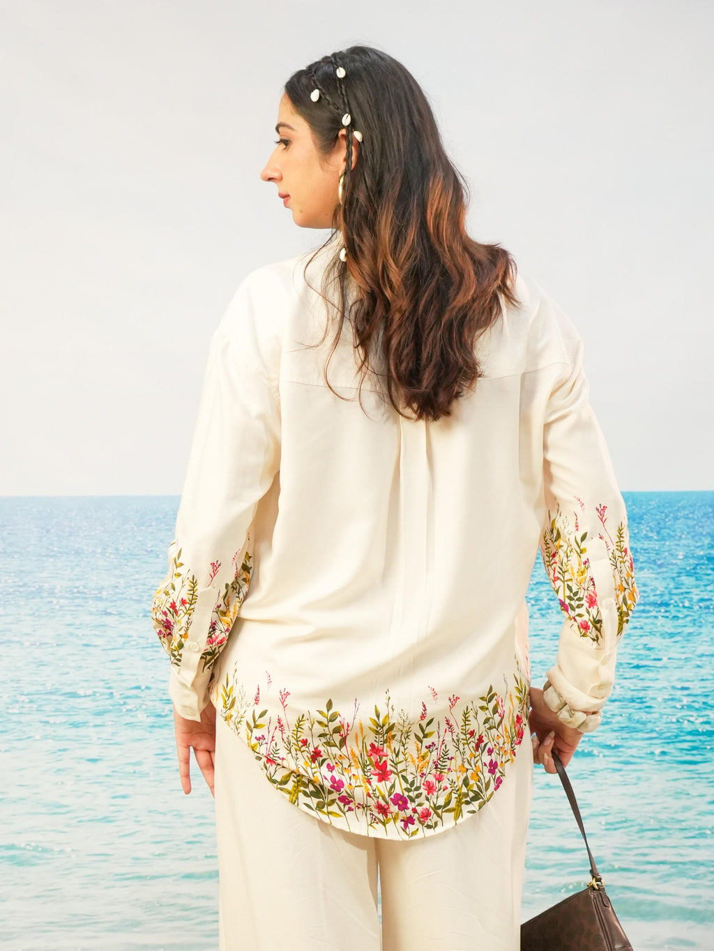A woman stands with her back to the camera wearing a cream-colored embroidered tunic top and matching trousers, with a tropical ocean background.