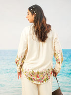 A woman stands with her back to the camera wearing a cream-colored embroidered tunic top and matching trousers, with a tropical ocean background.