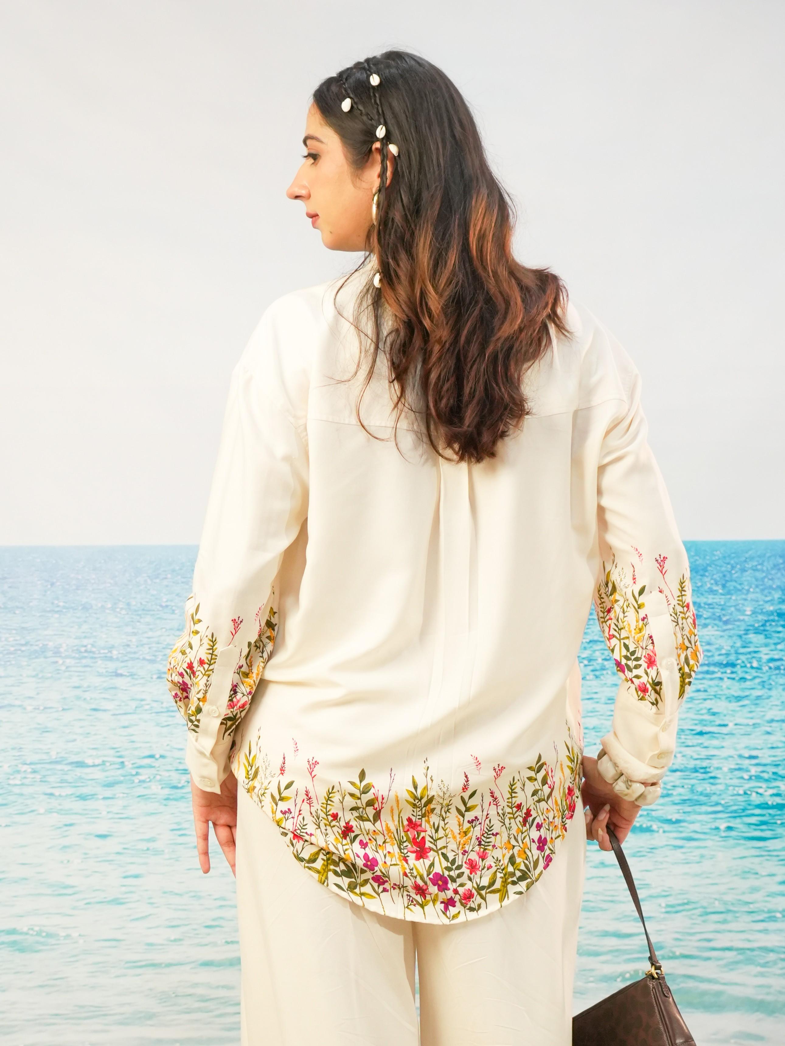 A woman stands with her back to the camera wearing a cream-colored embroidered tunic top and matching trousers, with a tropical ocean background.