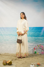 A woman stands on a beach backdrop wearing a long cream-colored embroidered kurta and wide-leg pants set, holding a brown handbag. The scene includes sand, ocean water, coconuts, and a colorful inflatable ring buoy.
