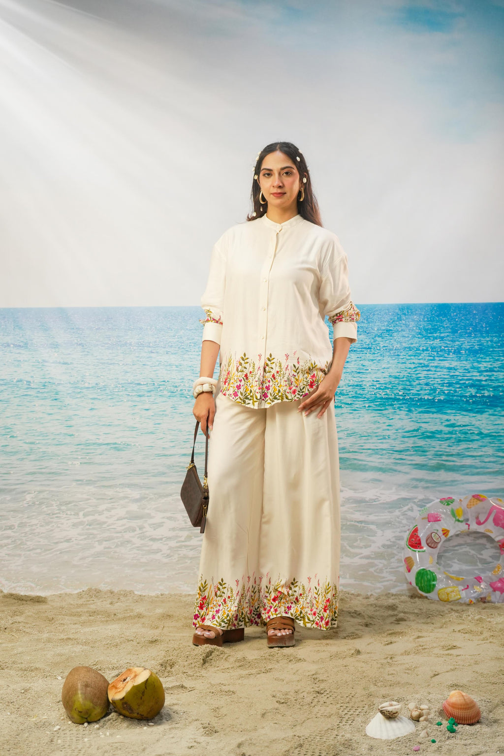 A woman stands on a beach backdrop wearing a cream-colored printed tunic and wide-leg pants set with floral detailing near the hem and sleeve cuffs. She holds a dark handbag, with coconuts and a colorful swim ring prop on the sand.