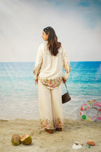 A woman stands on a sandy beach wearing a cream-colored printed tunic and wide-leg trousers set with colorful floral detailing. She is holding a dark brown handbag, with a blue ocean and sky backdrop, coconuts on the sand, and a colorful inflatable ring nearby.