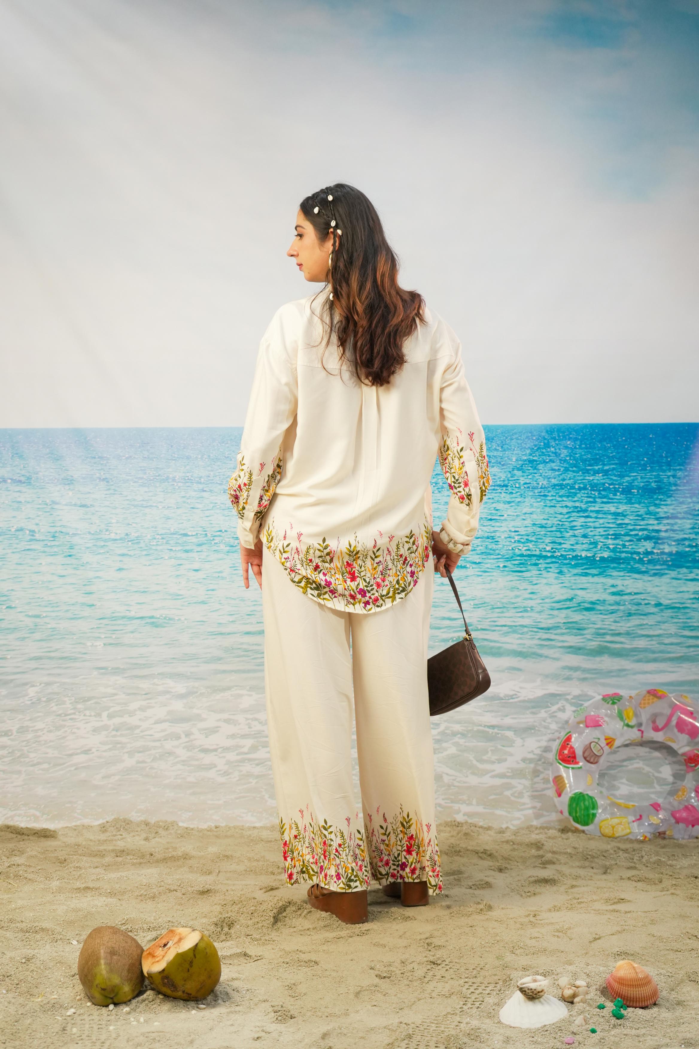 A woman stands on a sandy beach wearing a cream-colored printed tunic and wide-leg trousers set with colorful floral detailing. She is holding a dark brown handbag, with a blue ocean and sky backdrop, coconuts on the sand, and a colorful inflatable ring nearby.