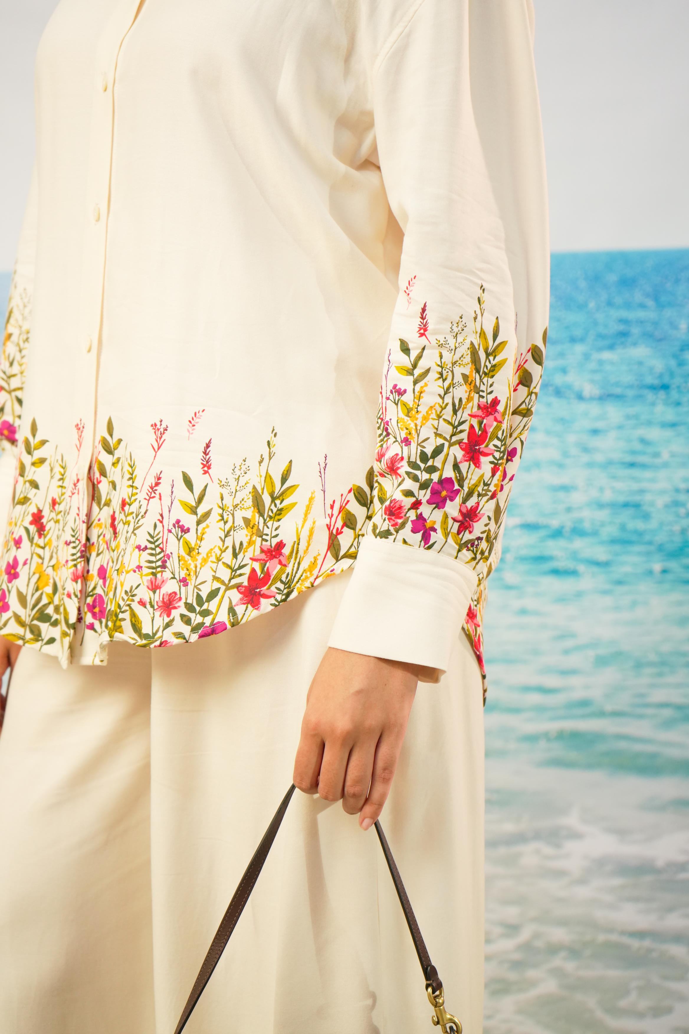 A close-up of a person wearing a cream-colored long-sleeve button-up shirt with colorful floral print around the hem and cuffs, paired with matching cream trousers, holding a black handbag strap against a beach background.