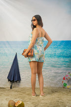 A woman stands on a beach backdrop wearing a light blue and white floral print romper, holding a brown clutch bag.