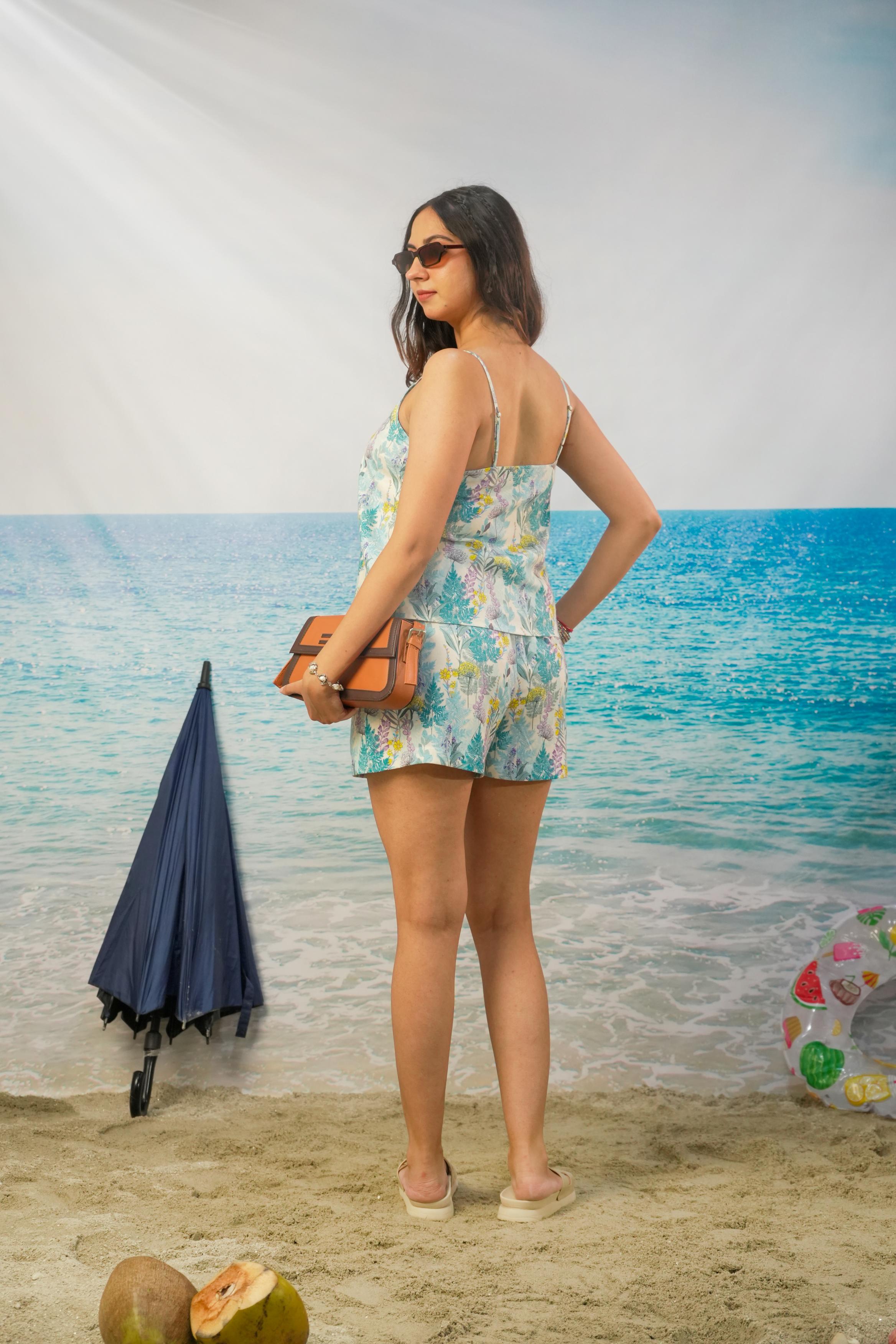 A woman stands on a beach backdrop wearing a light blue and white floral print romper, holding a brown clutch bag.