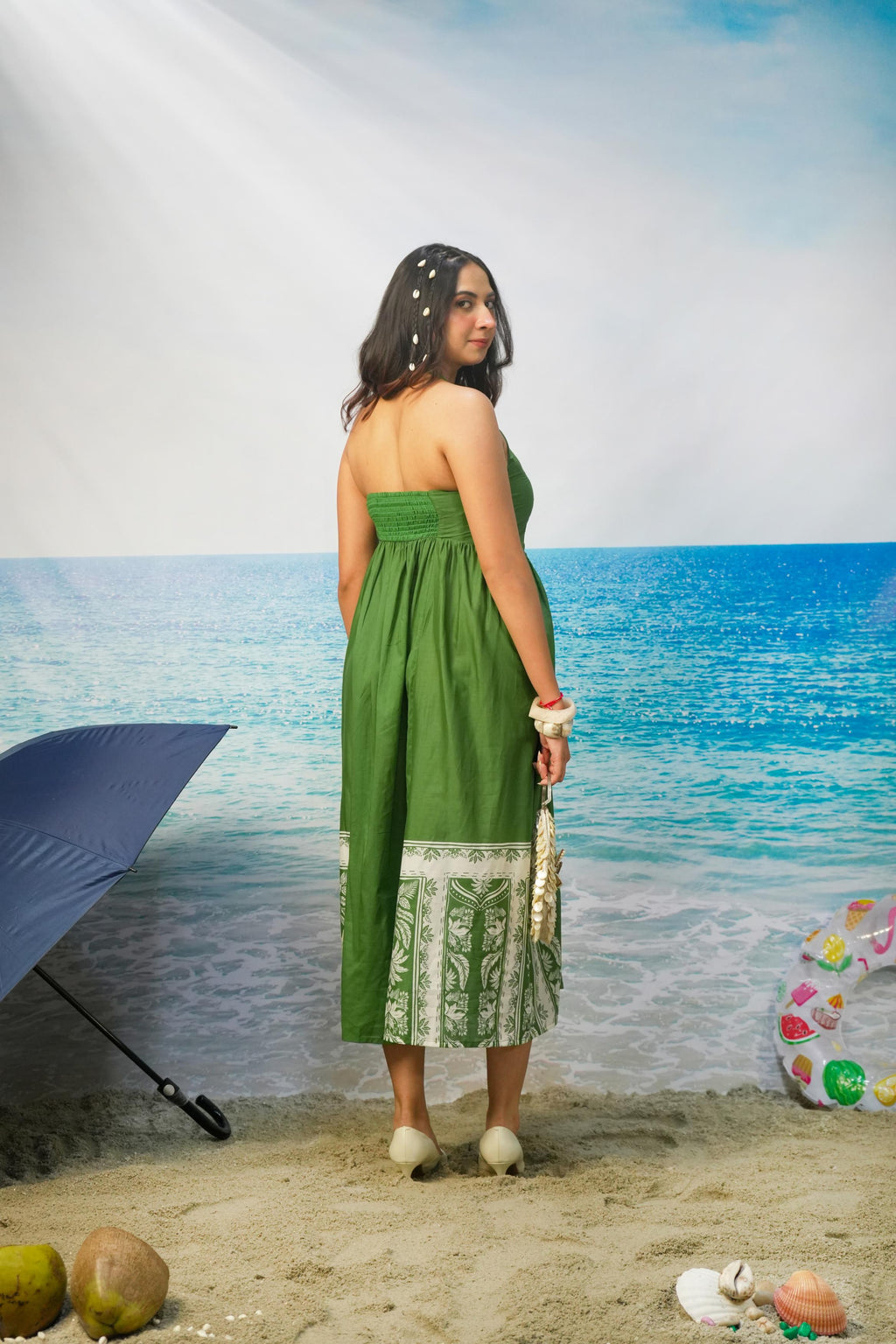 A woman stands on a beach backdrop wearing Tasneem Botanical Print Cotton Halter Neck Dress Back Image. She holds a small white handbag, with a blue umbrella, coconuts, and a colorful float visible around her.