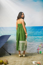 A woman stands on a beach backdrop wearing Tasneem Botanical Print Cotton Halter Neck Dress Back Image. She holds a small white handbag, with a blue umbrella, coconuts, and a colorful float visible around her.