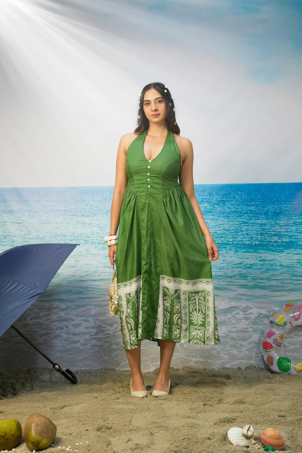 A woman stands on a beach backdrop wearing a Tasneem Botanical Print Cotton Halter Neck Dress with white printed border panels at the hem and white heels. The background depicts blue ocean water, sandy shore, and a blue umbrella at left.