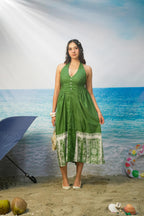 A woman stands on a beach backdrop wearing a Tasneem Botanical Print Cotton Halter Neck Dress with white printed border panels at the hem and white heels. The background depicts blue ocean water, sandy shore, and a blue umbrella at left.