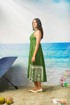A woman wearing aTasneem Botanical Print Cotton Halter Neck Dress on a sandy beach backdrop with the ocean behind her. A blue umbrella, coconuts, and a swim ring are placed around her.
