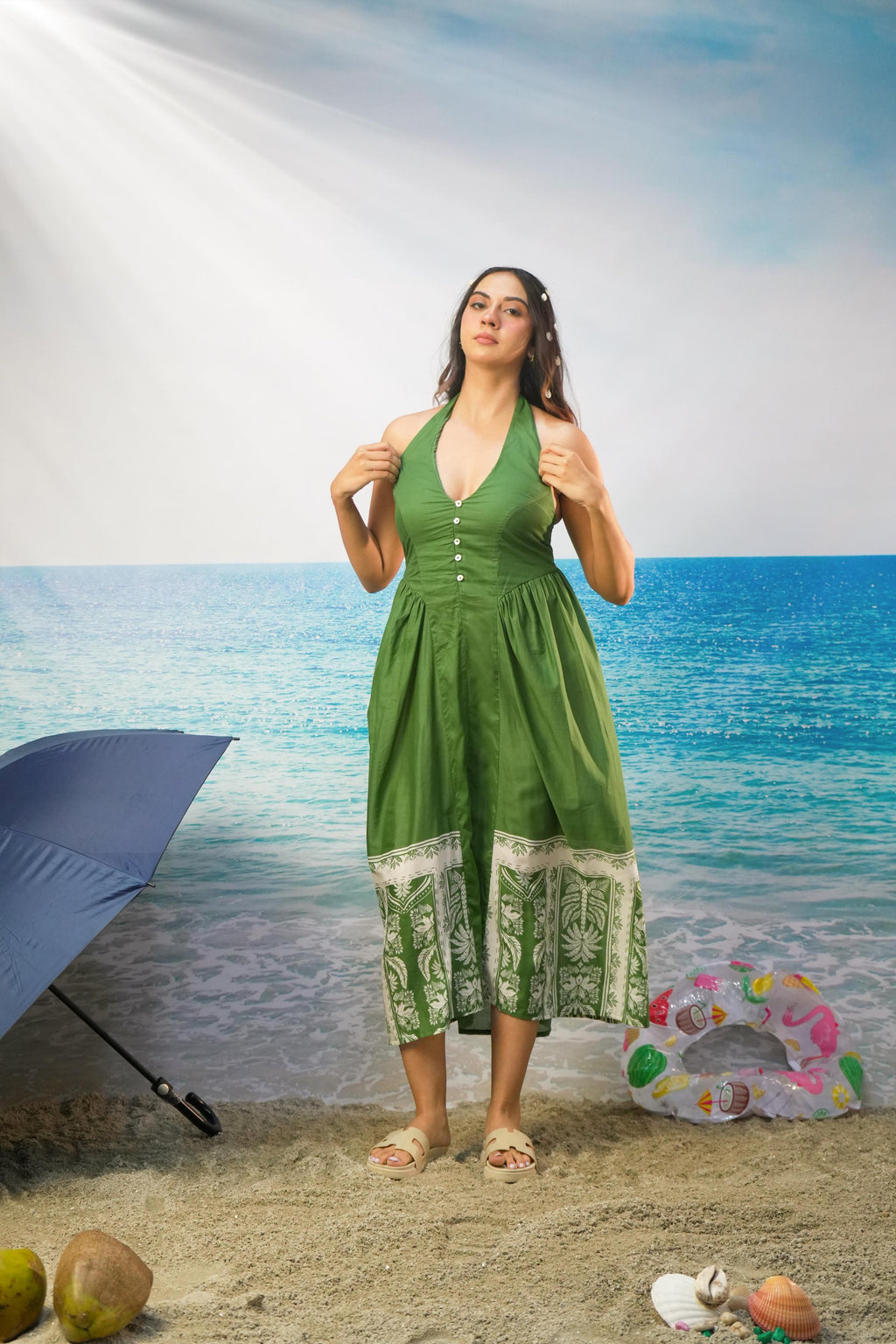 A woman modeling a Tasneem Botanical Print Cotton Halter Neck Dress with a white patterned hem, standing on a sandy beach backdrop with a blue umbrella, beach balls, and ocean scenery.