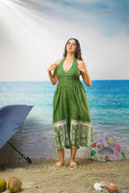 A woman modeling a Tasneem Botanical Print Cotton Halter Neck Dress with a white patterned hem, standing on a sandy beach backdrop with a blue umbrella, beach balls, and ocean scenery.