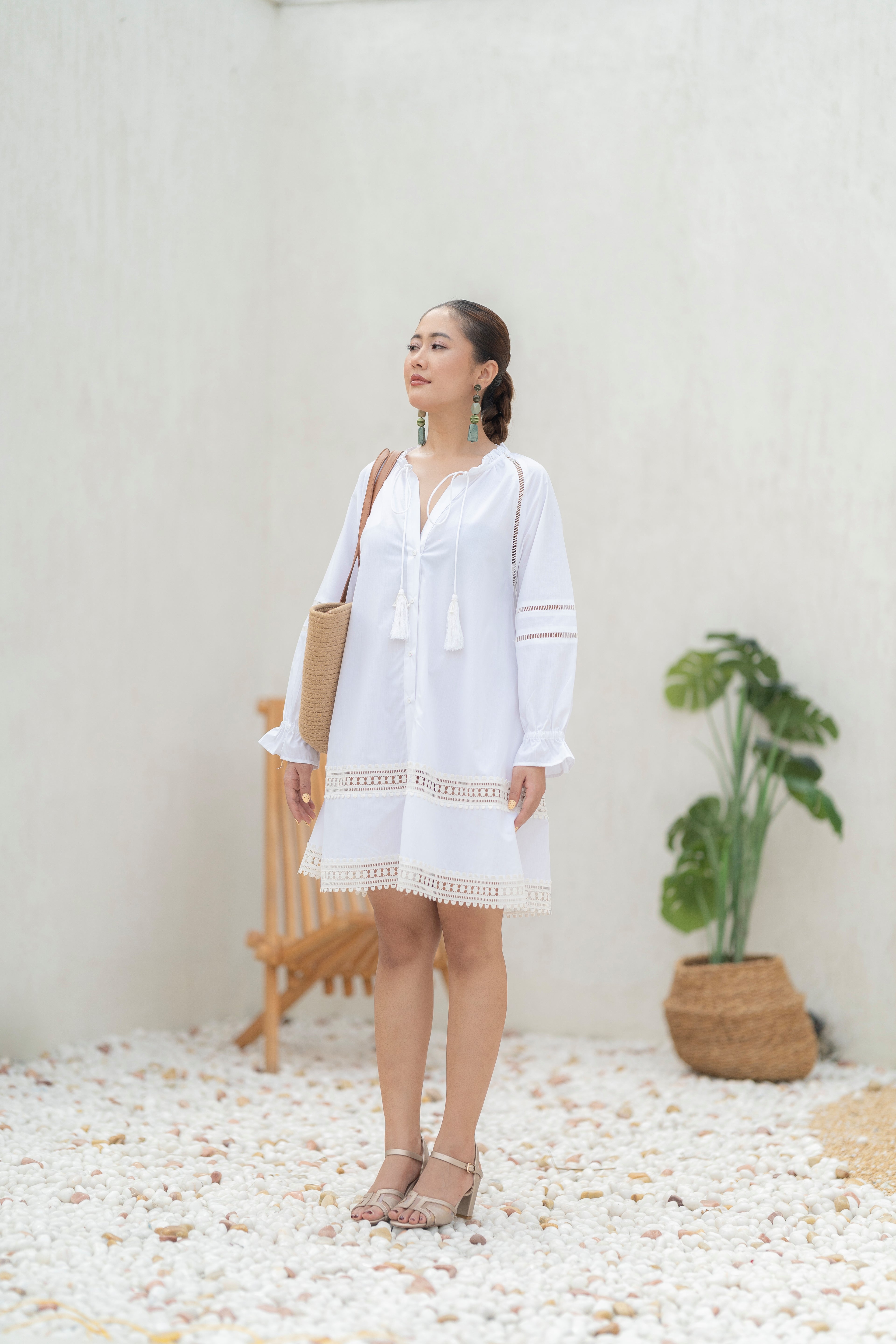 Woman in The Riviera Dress – White Cotton Poplin with Lace Detail standing on pebbles with a plant and chair in the background