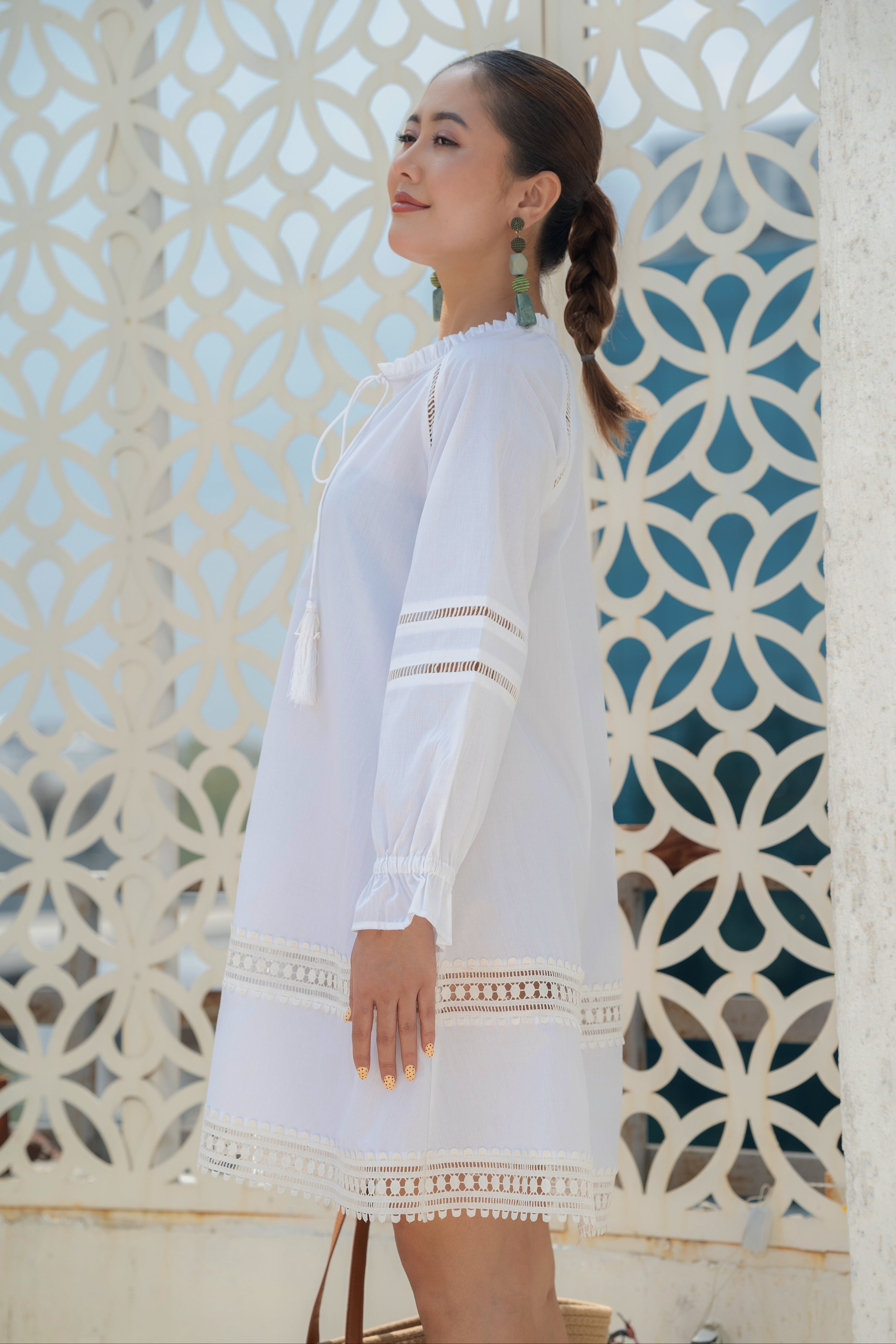 Woman wearing The Riviera Dress – White Cotton Poplin with Lace Detail with intricate patterns against a decorative white lattice.