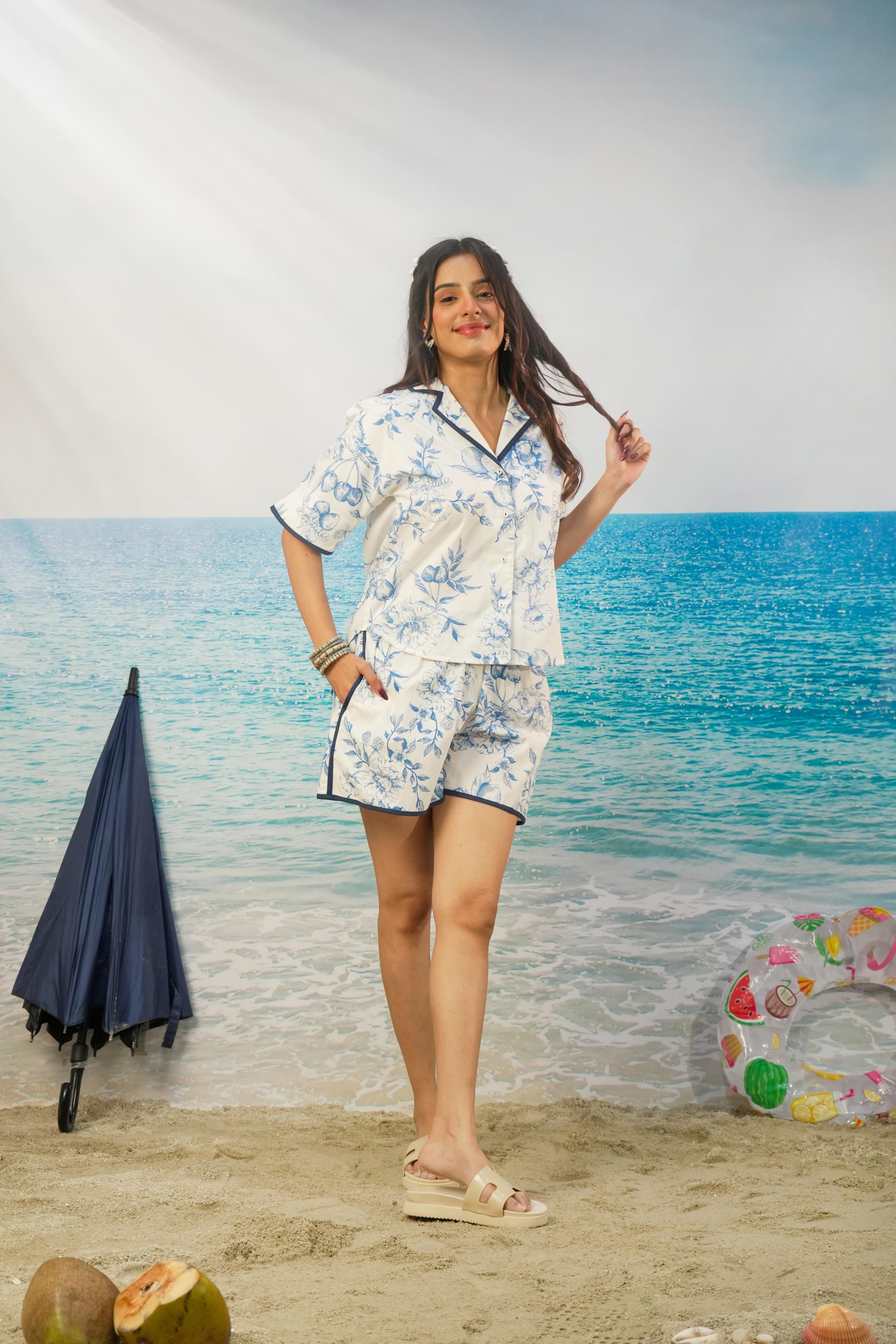 A woman modeling a matching two-piece pajama set with a blue and white floral print, standing on a beach-themed backdrop with ocean, sand, a closed umbrella, and a pool float visible.