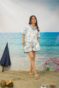 A woman modeling a two-piece pajama set with a blue tropical floral print on a white background, standing on a beach backdrop with a blue umbrella, coconuts, and a swim ring nearby.