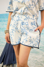 A person wearing a matching white two-piece set with a blue floral toile-style print and dark blue piping, standing against a beach backdrop with ocean water visible.