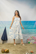 A woman modeling a white sleeveless midi dress with a blue floral print, standing on a sandy beach backdrop with ocean scenery behind her. Props include a closed navy umbrella, coconuts, and a colorful swim ring.
