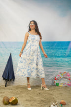 A woman modeling a white sleeveless midi dress with a blue floral print, standing on a sandy beach backdrop with ocean scenery behind her. Props include a closed navy umbrella, coconuts, and a colorful swim ring.