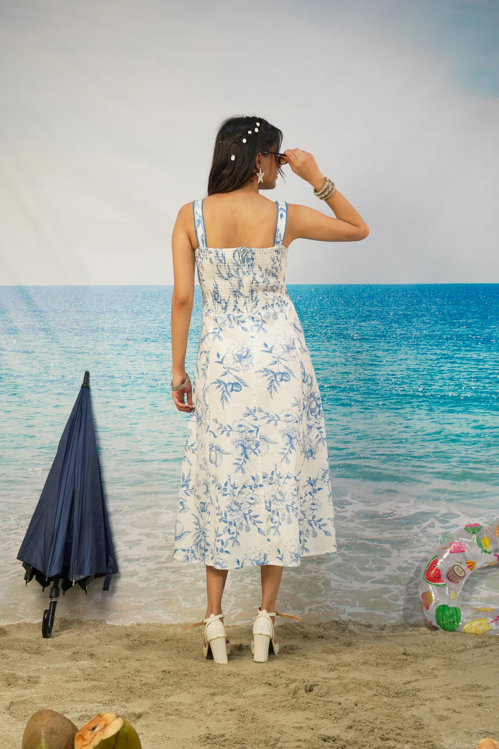 A woman stands on a beach backdrop wearing a white midi dress with a blue floral print, viewed from behind. The scene includes a blue umbrella, sand, ocean backdrop, and a colorful float.