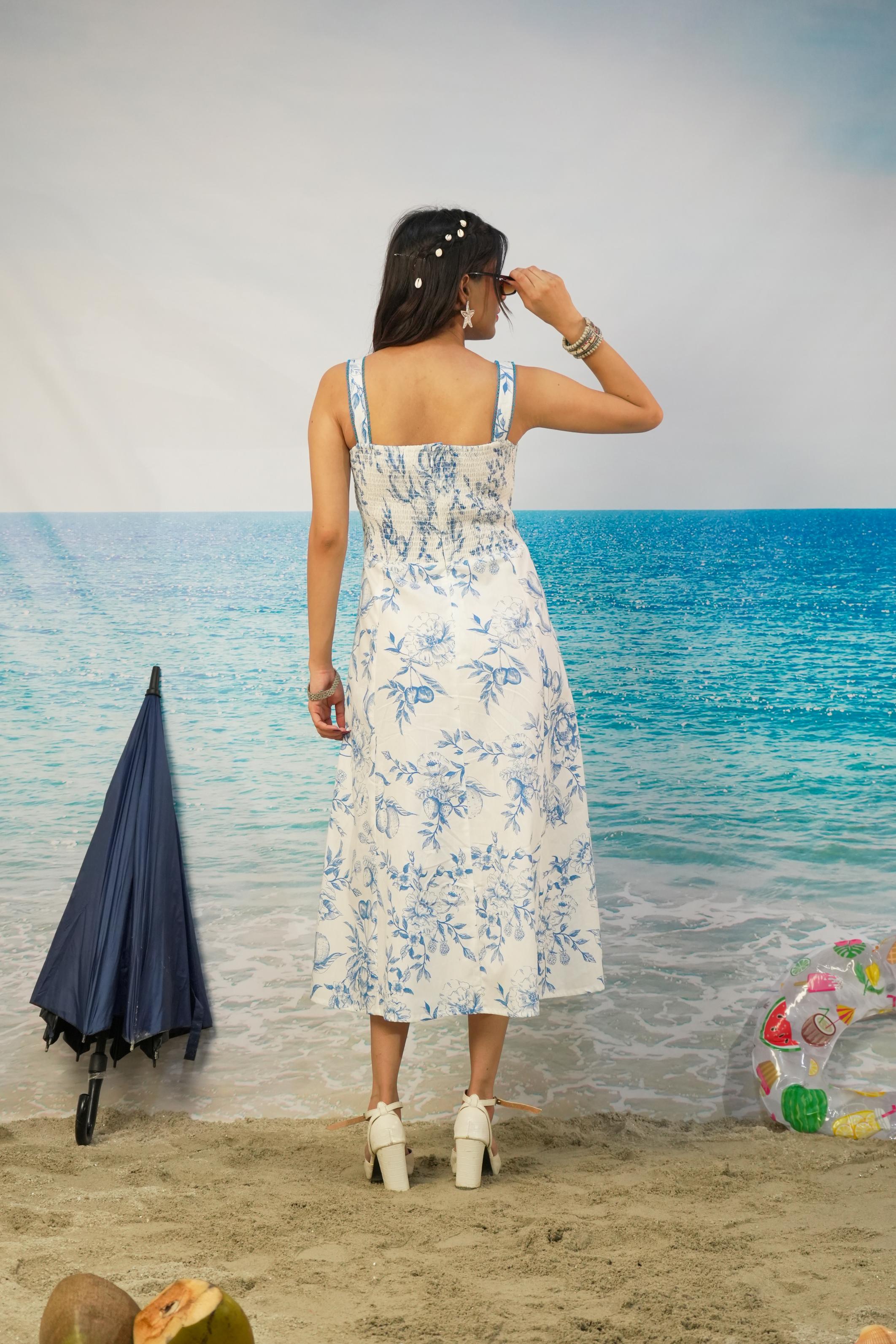 A woman stands on a beach backdrop wearing a white midi dress with a blue floral print, viewed from behind. The scene includes a blue umbrella, sand, ocean backdrop, and a colorful float.