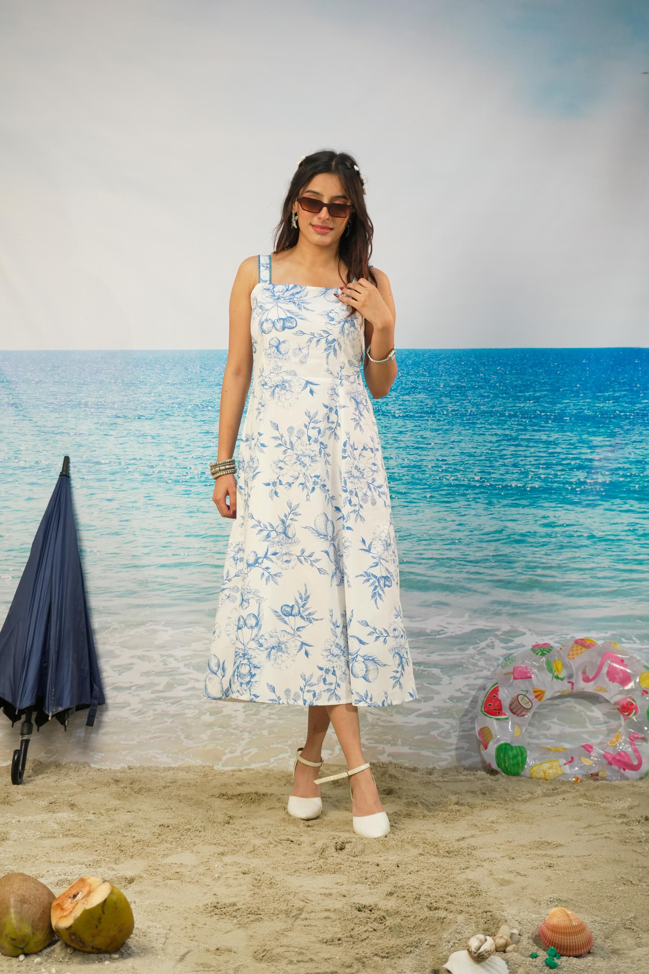 A woman modeling a white sleeveless midi dress with a blue floral pattern, standing on a sandy beach with a turquoise ocean backdrop. She is wearing white heeled sandals and sunglasses, with beach accessories like a closed navy umbrella, coconuts, and a pink swim ring nearby.