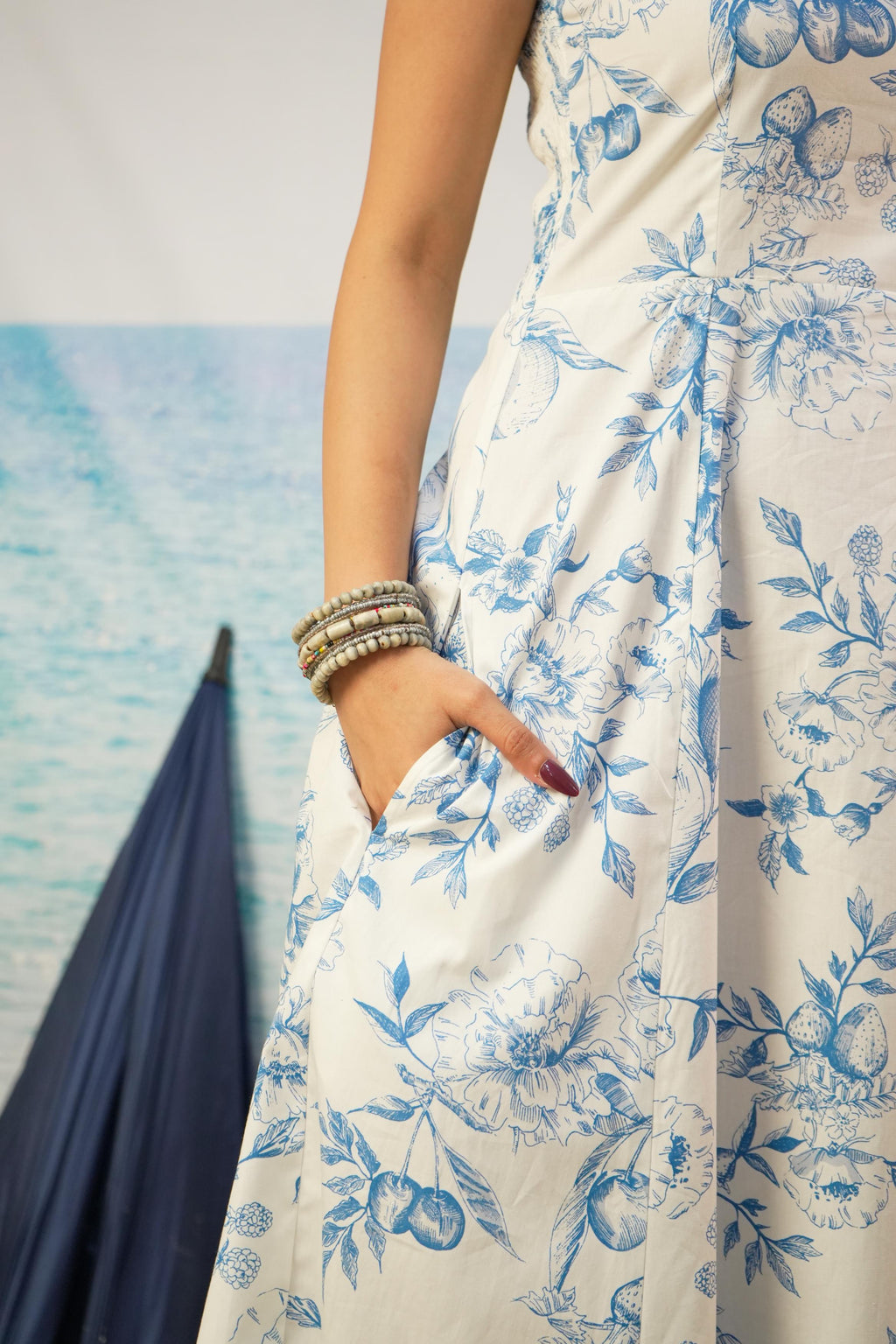 A close-up of a woman wearing a sleeveless white dress with a blue floral toile pattern, featuring pockets and a side slit, accessorized with stacked silver bracelets.